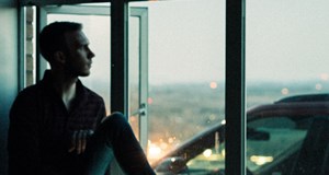 Man sitting on a window sill at dusk staring out of the window