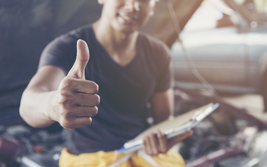 A person perched on an open car bonnet in a workshop smiling doing a thumbs up sign